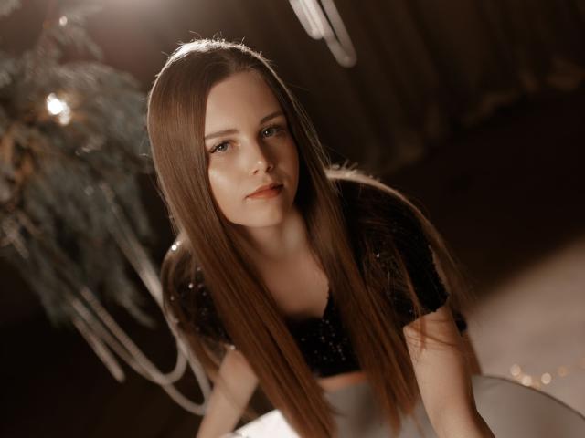 MileyGloss with long brown hair, wearing a black sparkly top, leaning forward, looking up with a slight smile, in soft, warm lighting, showing her tits and delicate features.