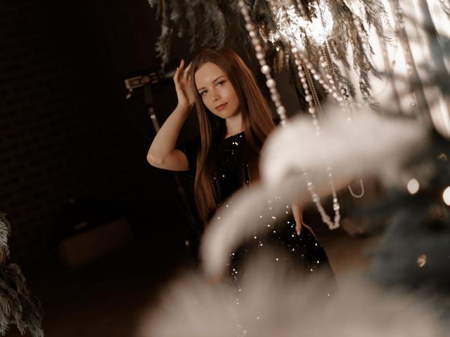 MileyGloss with long brown hair, wearing a black sequined dress, stands with one hand in her hair, looking softly at the camera. Her tits are partially visible.