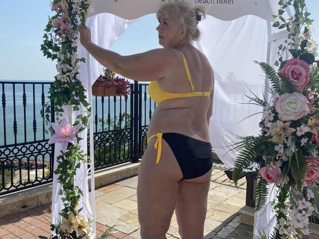 BozhenaNude, kurvig mit heller Haut und blonden Haaren, trägt einen gelben BH und schwarze Bikini-Hosen und steht unter einem floralen Hochzeitbogen, mit dem Rücken zum Betrachter, wobei ihr Hintern zu sehen ist..