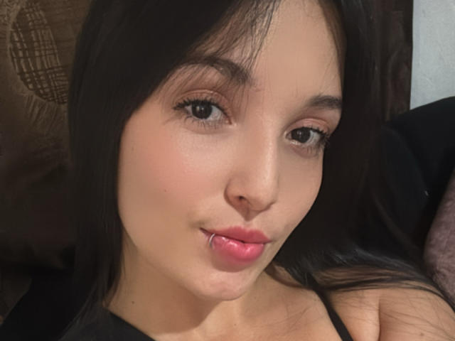 SarahGood has light brown skin, dark brown hair, and a small nose piercing on her lower lip. She's looking at the camera with pink lipstick and black eyes.