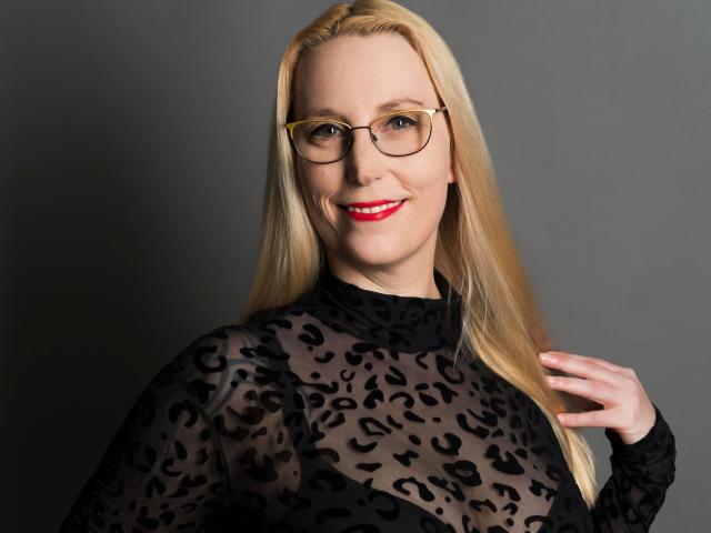 SallySecret-hot with blonde hair, glasses, and red lipstick, wearing a sheer black leopard print top, smiling, hand near neck.