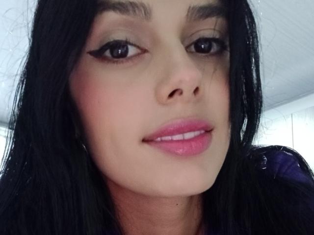 Anais Hilton tiene el cabello negro, lápiz labial rosa y delineador de ojos, con una ligera sonrisa, mostrando sus labios carnosos y seductores. Está mirando a la cámara con ojos marrones..