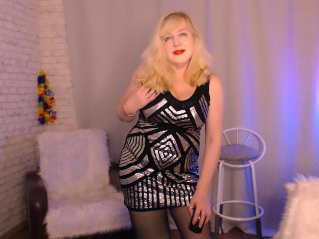 SamanthaLala with blonde hair, blue eyes, red lipstick, wearing a black and silver geometric dress, standing, hand on hip, white brick wall, chair, and stool in background.