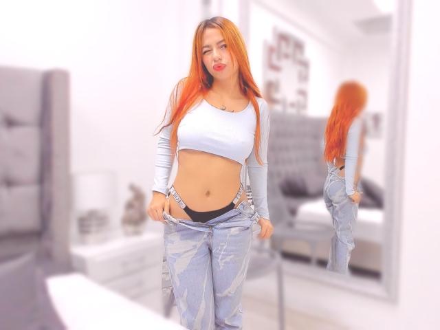 Samanta Carter, con lunghi capelli arancioni, indossa un top bianco a vita corta che lascia scoperto l'addome, e tira giù i jeans blu per rivelare delle mutande nere, mentre è in piedi di fronte a uno specchio..