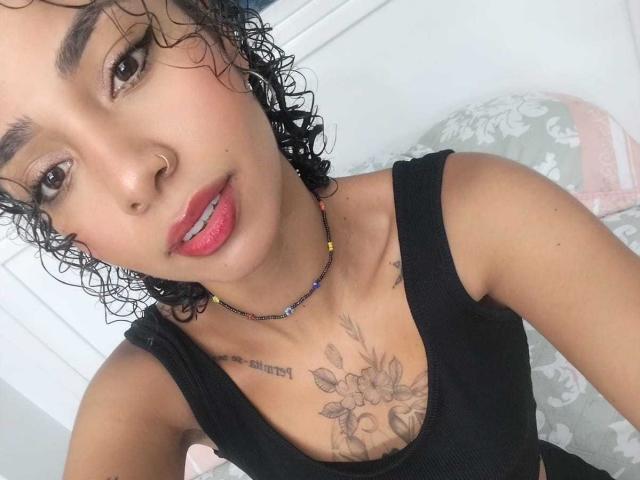SilvanaXOwen with curly black hair, light brown skin, pink lipstick, black tank top, floral chest tattoo, beaded necklace, and a subtle smile. Her tits are visible under the top.