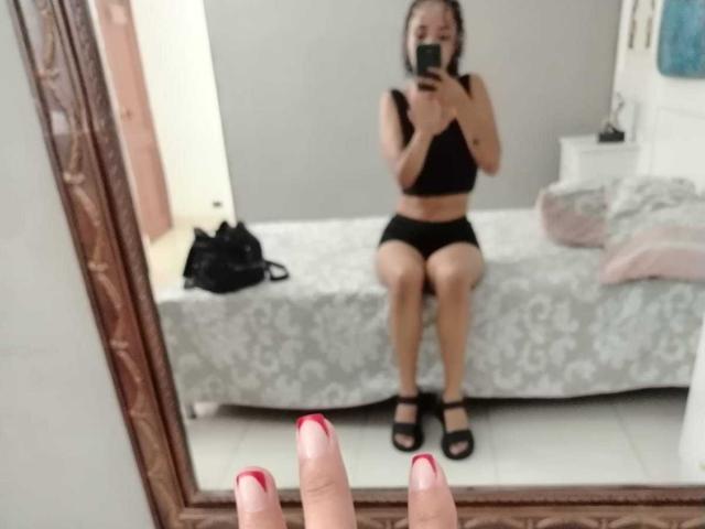 SilvanaXOwen, met kleine borsten, draagt een zwarte bh en korte broek en zit op een bed. Ze maakt een spiegelzelfportret. Haar zwarte schoenen en rode nagels zijn in beeld..