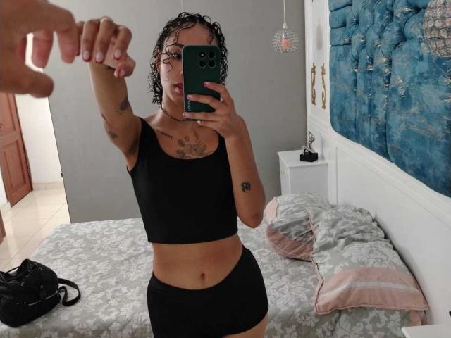 SilvanaXOwen, with wet curly hair, takes a mirror selfie in a black sports bra and shorts, showing tattoos and a chest tattoo, holding her phone with a hand in frame.