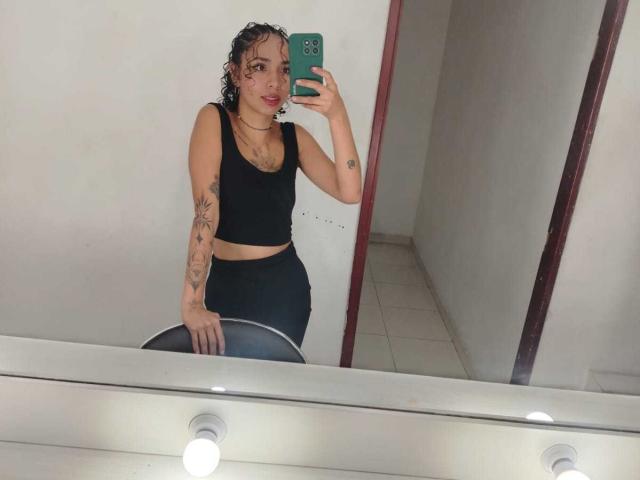 SilvanaXOwen, con i capelli castano ricci, rossetto rosso, reggiseno sportivo nero e pantaloncini neri, tiene in mano un telefono verde, mostrando tatuaggi sul braccio destro, ed è in piedi di fronte a uno specchio..