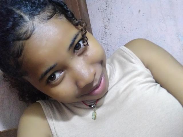 SexyEmilyStar69 with curly black hair, medium brown skin, and a cute smile, wearing a beige sleeveless top, has a small green pendant necklace, and is looking at the camera.