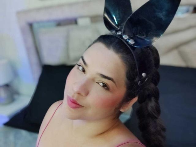 AntonelaPark with dark hair in a braid, wearing black shiny bunny ears with pearls, pink spaghetti straps, and pink lipstick, looking up with a cute pout.