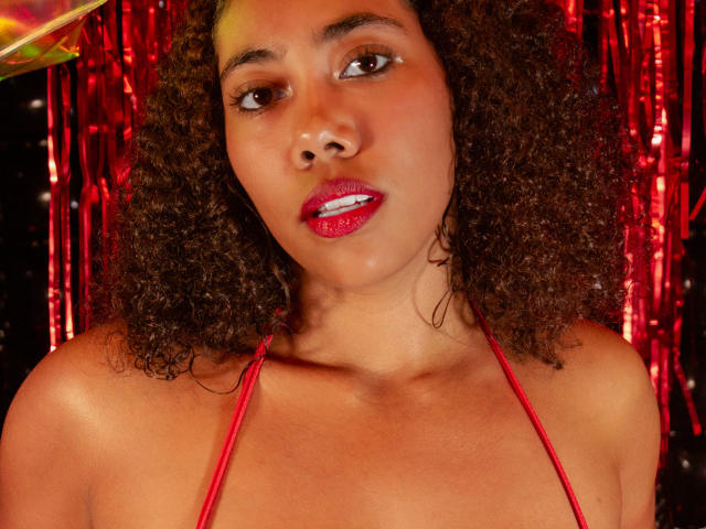 AishaRhodes with curly brown hair, red lipstick, and red halter top, showing her tits, against a red, shiny background. Her brown eyes look slightly to the side.