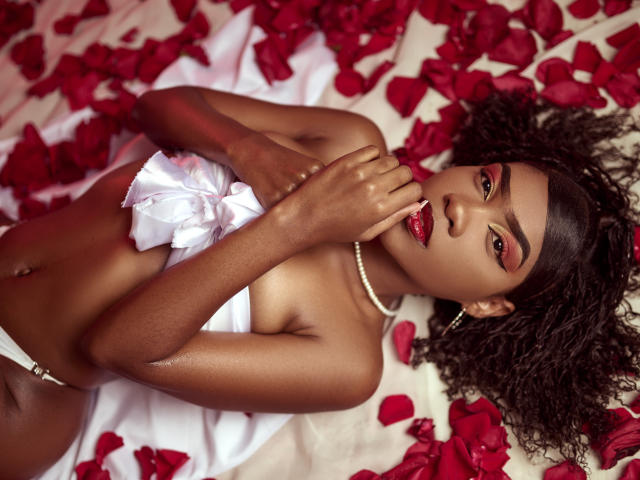 Alana Patel, mit dunkler Haut, lockigem Haar, rotem Lippenstift, weißem Band auf der Brust, einer Perlenkette, liegt auf roten Rosenblättern, der Finger ist in der Nähe des Mundes, sie schaut in die Kamera, sexy..