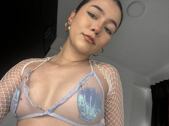 DeisyWest, con i capelli scuri raccolti in uno chignon, indossa una trasparente maglia a rete bianca e un reggiseno a fascia blu metallizzato, mostrando piccoli seni con capezzoli visibili, grandi orecchini a cerchio e un leggero sorriso..