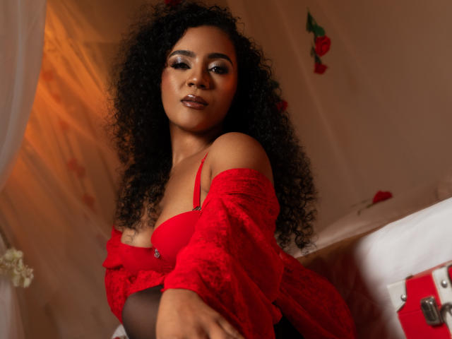 PaolitaKens with curly black hair, red bra, and sheer red top, showing her tits, sitting on a bed, looking at the camera with a sultry expression.
