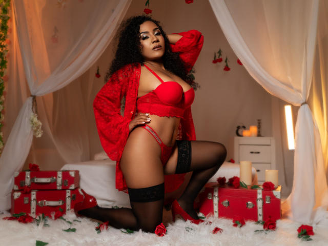 PaolitaKens with curly hair, red lingerie, and lace robe, kneeling, showing her tits and pussy, wearing black stockings, on a white fur rug with roses and red trunks.