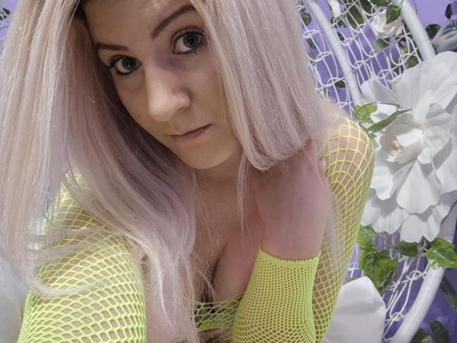 HoneyPearl with blonde hair, green eyes, and a cute face, wearing a neon yellow fishnet top, showing her boobs, leaning forward with a hand near her mouth.