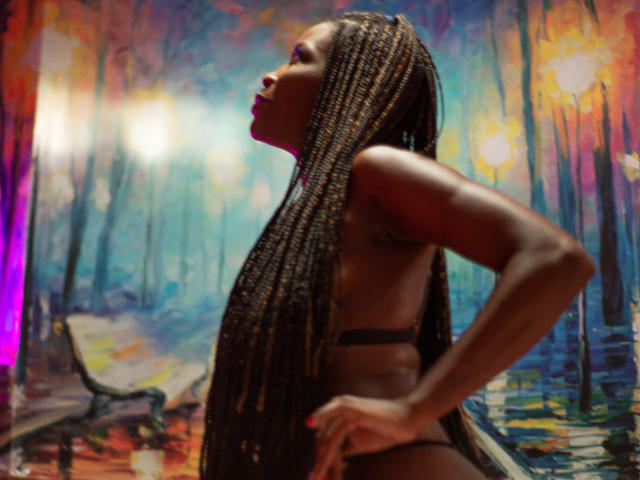 MaxAndMegan69, dark-skinned, topless with long braided hair, hands on hips, facing side, in a colorful, forest-like backdrop, glowing lights on left.