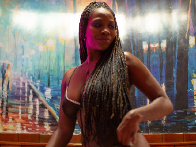 MaxAndMegan69, dark-skinned woman with long braids, small tits, wearing sheer black top, pink and blue eye makeup, smiling, sitting in front of colorful, abstract background.