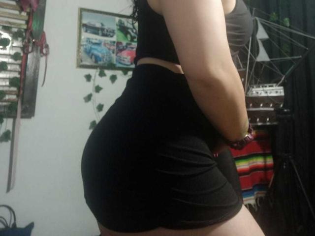 CharloteEzra with a curvy, round ass in tight black shorts and a black crop top, her arm down, wearing a red watch, in a dimly lit room.
