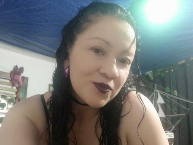  CharloteEzra has wet black hair, dark purple lipstick, and a black choker. She's topless, with visible shoulders and a slight smile. A bright light is in the top right.