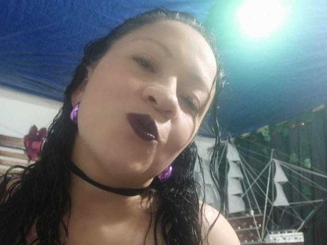 CharloteEzra with wet black hair, purple earrings, black choker, and dark purple lipstick, making a pouty face, under bright green light.