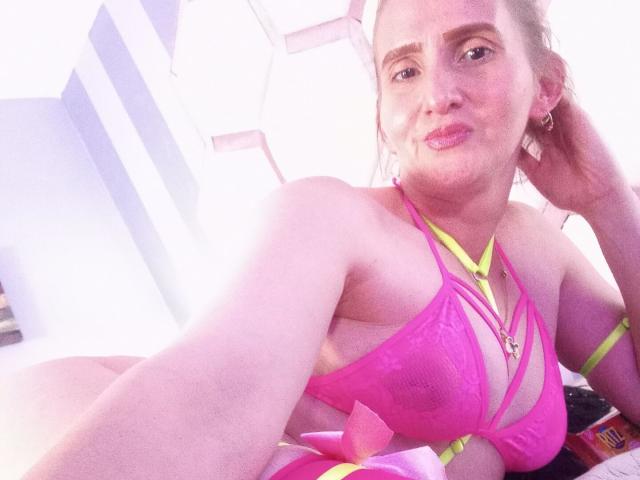 Rubi Anderson usando um biquíni rosa com alças neon, mostrando os seios, deitada, fazendo beicinho com os lábios rosados, cabelo loiro, pele clara, braço direito levantado, luz solar forte..