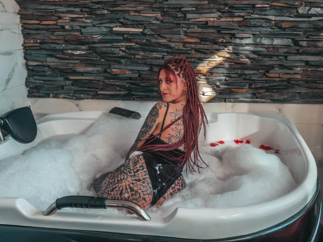SofiFw in a bubble-filled hot tub, looking back over her shoulder with red braided hair, black wet dress, and tattooed back, surrounded by foam with red rose petals.