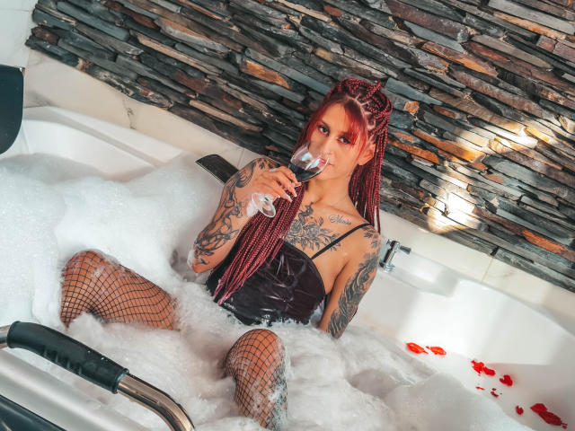 SofiFw with red braids, tattoos, fishnet legs, black velvet top, sipping wine in a bubble-filled tub, stone wall in background, holding glass in right hand.