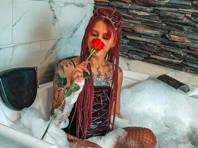 SofiFw with red braids, tattoos, and fishnet stockings, holding a rose to her mouth, sitting in a bubbly bathtub, wearing a black corset, looking down.