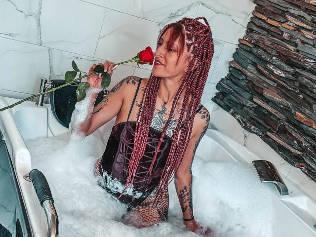 SofiFw with pink braids, holding a red rose in a bubble-filled bathtub, wearing a black corset, showing tattoos, and a necklace, looking sexy and relaxed.