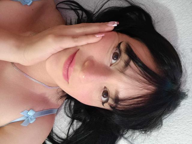 VaaleryStark has black hair, brown eyes, and pink lips, lying down with a hand covering part of her face. She's wearing a blue strap with a bow.