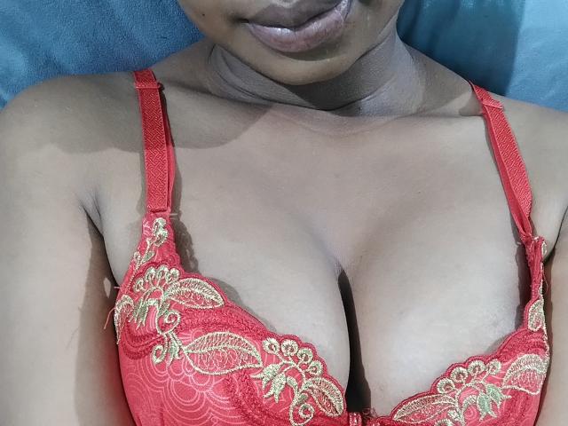 Avika's dark brown skin and red lace bra with gold embroidery highlight her large, round boobs. Her full lips are slightly parted. She's lying down.