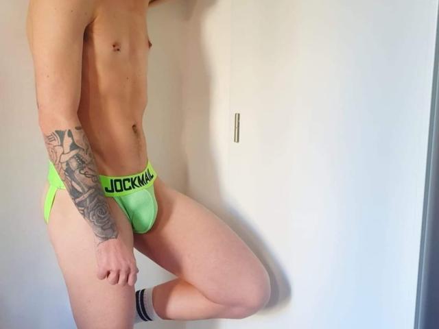 AlessRicalle in neon green underwear, showing a tattooed arm, white socks, and a lean, muscular body, standing with one leg up. No visible genitals.