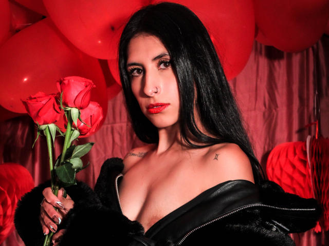 BondageGirlKs with long black hair, red lipstick, and tattoos, holding red roses, wearing a black leather jacket, showing her tits, in a red balloon-filled setting.