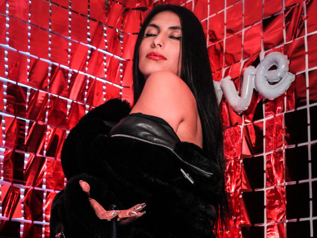 BondageGirlKs with long black hair, red lipstick, and black leather top, wearing black fur jacket, eyes closed, smiling, red background, and white balloon letters.