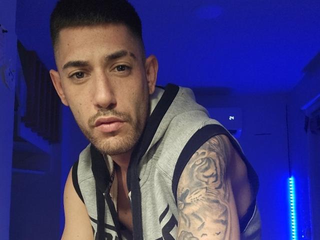 AlessRicalle with short dark hair, light brown skin, and a tattooed arm wearing a sleeveless gray hoodie, looking into the camera with a serious expression. Blue room lighting.