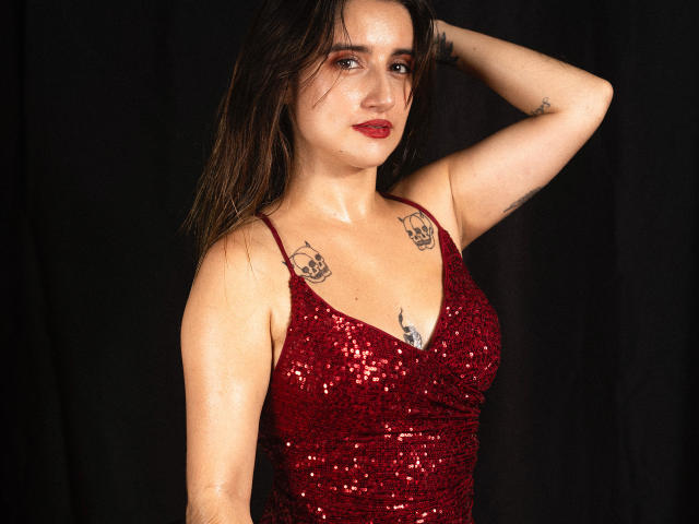 LindaSpark con un vestido rojo brillante adornado con lentejuelas, con una mano en el pelo, tatuajes en el pecho, lápiz labial rojo y vello axilar visible, de pie frente a un fondo oscuro. Se ven los senos, los tatuajes y el vello de los brazos..