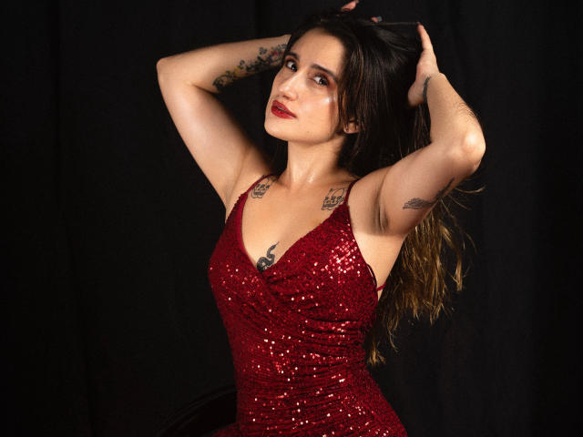 LindaSpark with arms raised, wearing a red sequined dress, showing tattoos on arms and chest, long brown hair, red lipstick, black background. Boobs, tits, armpits, arms, chest tattoos.