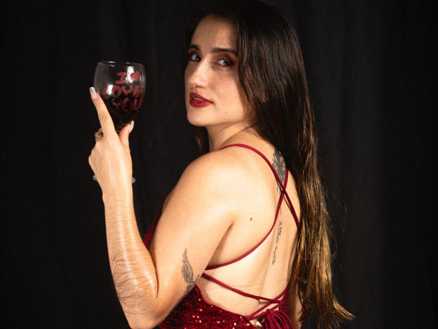 LindaSpark with long brown hair, red dress, holding wine glass, tattoos on arm and back, looking over shoulder, red lipstick, dark background, right arm raised, medium shot.