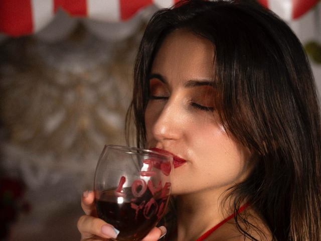 LindaSpark, con cabello oscuro, ojos cerrados, lápiz labial rojo, sosteniendo una copa de vino tinto, con los labios rozando suavemente el borde, una luz suave en su rostro, un brillo sutil en su piel..