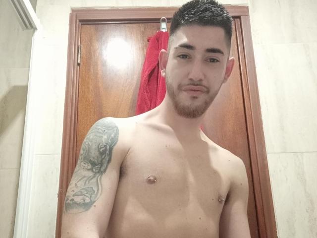 AlessRicalle, shirtless with a tattoo on his left shoulder, short black hair, and a beard, has a slight smile, visible nipple, and a red towel behind a wooden door.