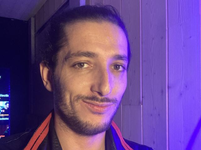 Malougaytrav has a medium skin tone, dark hair in a ponytail, a beard, and a slight smile. His face is illuminated by blue and purple lights.