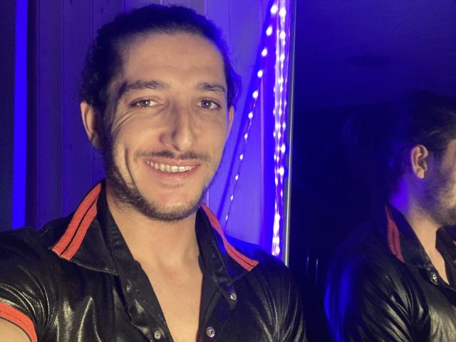 Malougaytrav with a slight grin, wearing a shiny black leather shirt with red trim, has a short beard, and dark curly hair, in a blue-lit setting.
