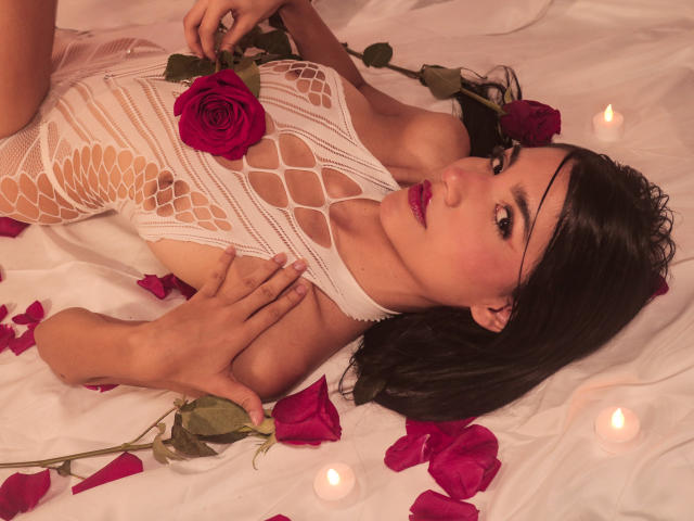 RoxannaHerias está deitada em lençóis brancos, vestindo um vestido de renda branca com detalhes de peixe, com rosas e velas. Ela tem batom rosa, cabelo escuro, e suas mãos estão sobre os seios..