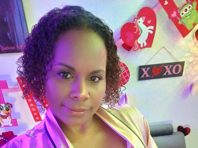 AfroditaKane, con cabello negro rizado, piel marrón media y senos pequeños, vestida con una camisa rosa, sonriendo ligeramente, en una habitación con coloridas decoraciones de San Valentín..