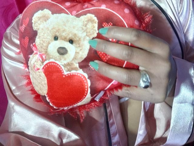 AfroditaKane's hand with mint green nails holds a red heart-shaped pillow with a stuffed beige teddy bear, wearing a shiny pink satin dress, silver ring on index finger.
