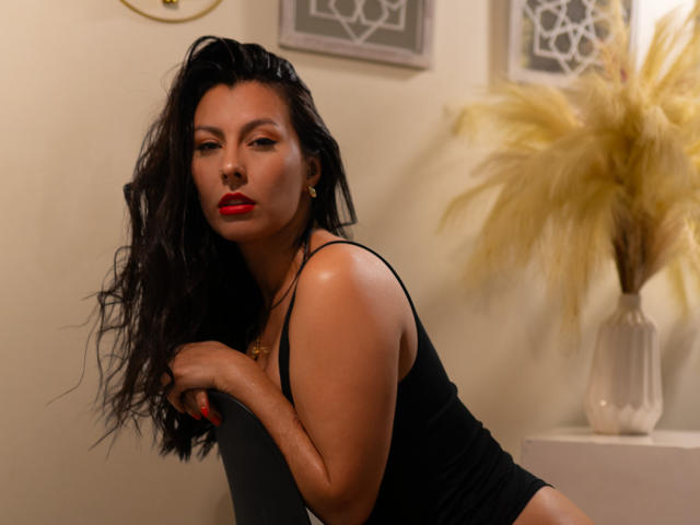 AnnyCraffort with long black hair, red lipstick, black tank top, and gold earrings, leaning on a black chair, looking seductively at the camera. Her tits are slightly visible.