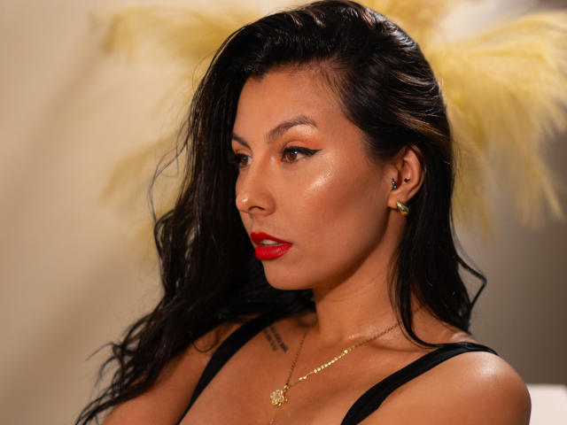 AnnyCraffort has glossy brown skin, dark wavy hair, red lipstick, gold earrings, and a necklace with a star charm, showing off her tits in a black top.