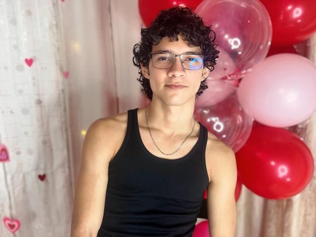 SamuelHills with curly black hair, glasses, and a black tank top, wearing a silver necklace, standing in front of red and pink balloons, smiling subtly.