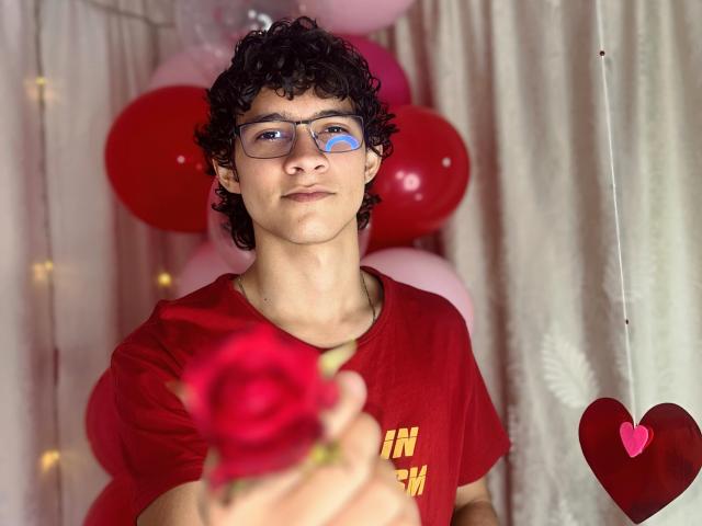 SamuelHills, met krullend zwart haar en een bril, houdt een rode roos vast, draagt een rood shirt en kijkt met een lichte glimlach in de camera. Op de achtergrond zijn er roze en rode ballonnen..